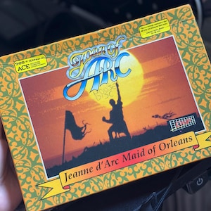May include: A rectangular video game box for Joan of Arc: Jeanne d'Arc Maid of Orleans. The orange border features a green pattern. The center illustration shows a silhouette against a yellow sun, with the handwritten text MOOSE DISC on the front.