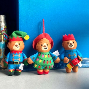 Può includere: Tre ornamenti natalizi di peluche a forma di orso. L'orso a sinistra indossa un cappello da elfo verde e un vestito blu e rosso. L'orso al centro indossa un cappello rosso e un vestito verde con luci bianche. L'orso a destra indossa un cappello rosso e un cappotto blu con un regalo rosso e giallo.
