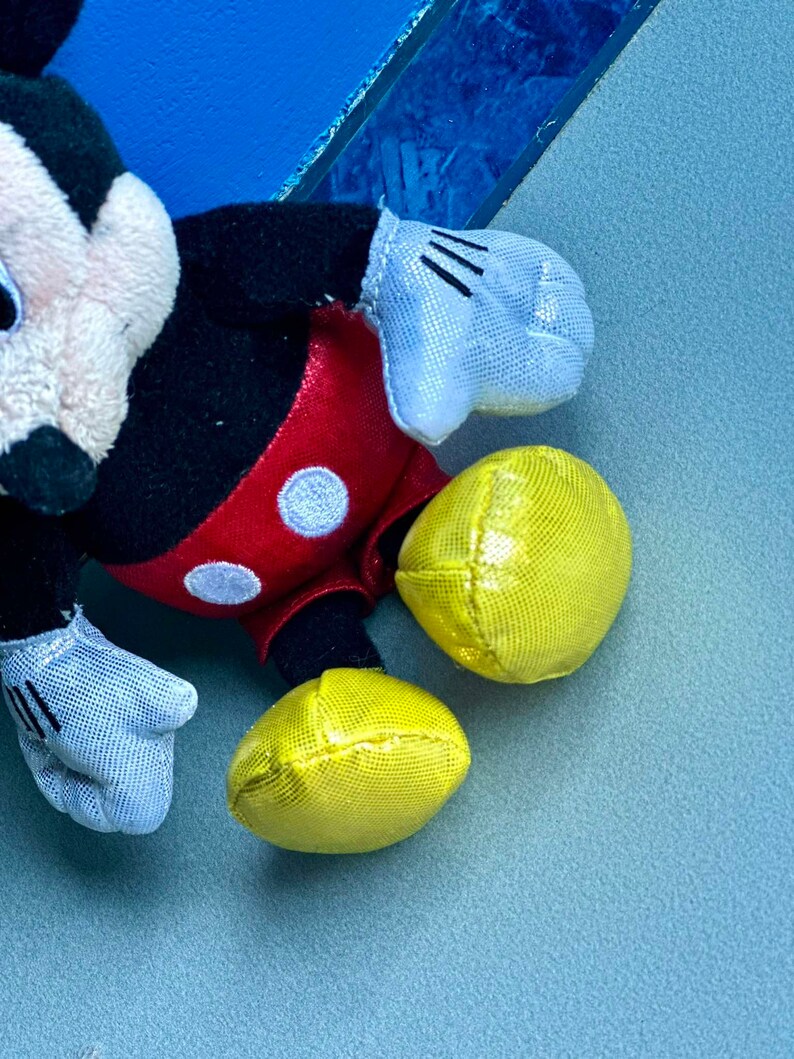 May include: A black and red plush Mickey Mouse toy with yellow ears and white gloves. The toy is lying on a blue surface.