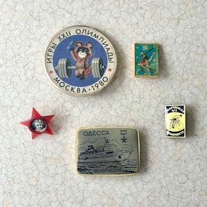 May include: A collection of vintage enamel pins featuring a red star with a portrait, a red, white, and blue pin with a bear lifting a barbell, a blue and white pin with a ship, a blue and white pin with a figure skater, and a yellow and blue pin with a cyclist.