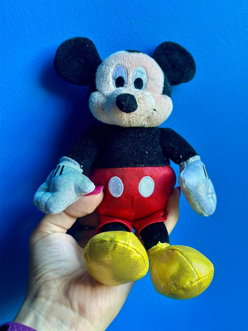 May include: A plush Mickey Mouse toy with black fur, large ears, and a red and white outfit. The toy has a yellow mesh fabric on the shoes.