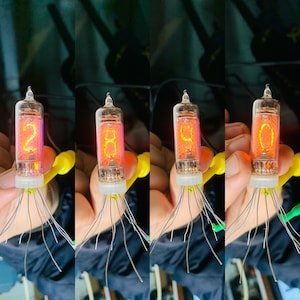 Peut inclure: Quatre tubes Nixie en verre transparent avec des chiffres orange brillants 2, 8, 9 et 0. Chaque tube possède de nombreux fils métalliques fins qui s'étendent du bas.