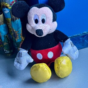 May include: A plush Mickey Mouse toy with black ears, a black body, red shorts with two white circles, and yellow shoes. The toy is sitting on a gray surface.