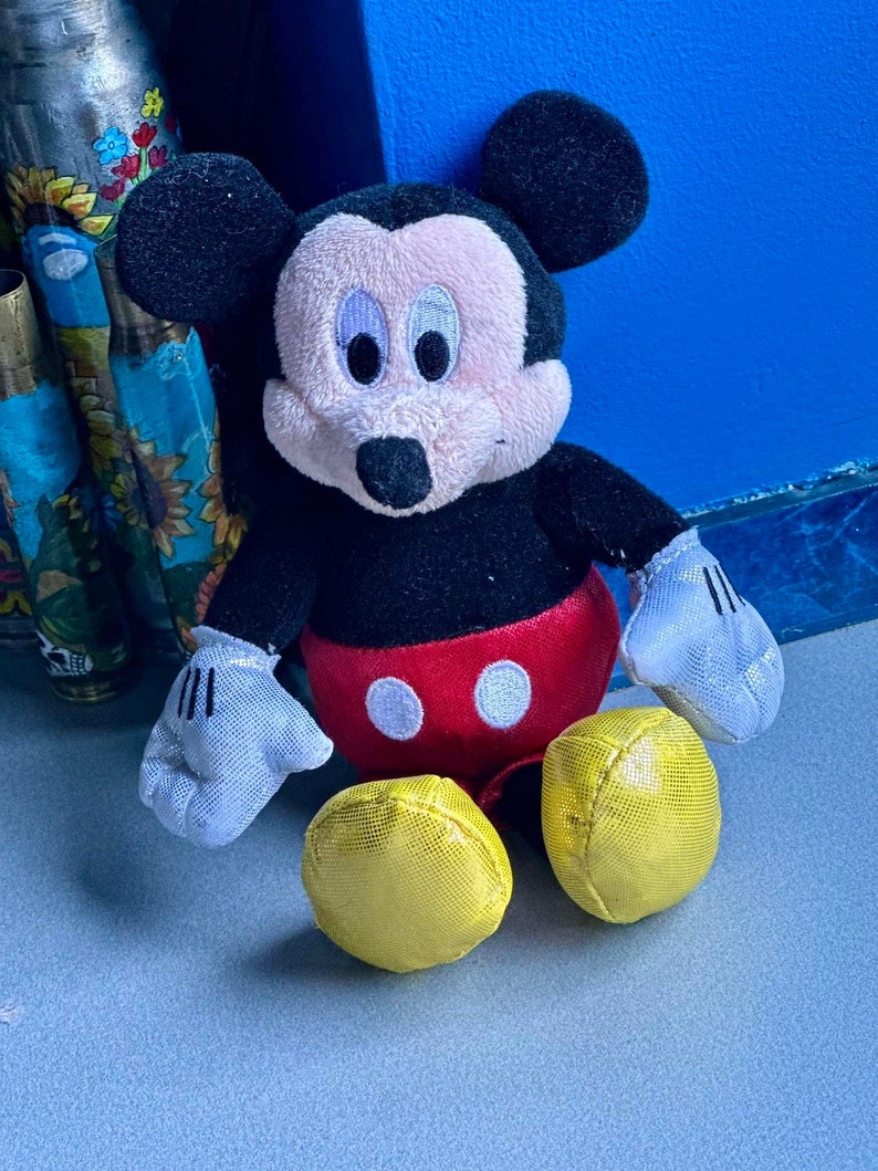 May include: A plush Mickey Mouse toy with black ears, a black body, red shorts with two white circles, and yellow shoes. The toy is sitting on a gray surface.