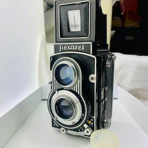 May include: A black and silver Flexaret medium format camera with a light meter on top. The camera has a lens with a silver ring and a black leather strap.