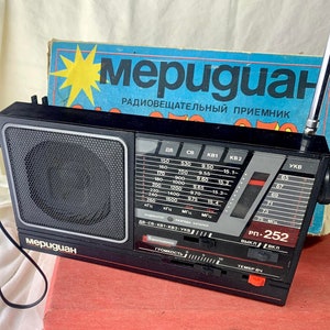 Peut inclure: Une radio AM/FM noire et argentée avec une grille de haut-parleur noire et un bouton rouge. La radio possède un écran numérique avec des marques de fréquence et un cadran pour la mise au point. La radio est étiquetée "меридиан" et "РП-252".