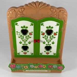 May include: A vintage toy cabinet in shades of brown, green, and white. The cabinet features two doors with apple-shaped cutouts and decorative floral and heart accents. The base has a floral border, and the text "Life is Berry Delicious" is visible.