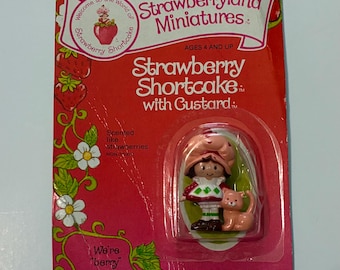 May include: A vintage Strawberry Shortcake miniature figure with custard, packaged on a red and pink card. The card features the text "Strawberryland Miniatures" and "We're berry happy together." The figure is scented like strawberries.