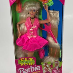 May include: A Barbie doll in a pink dress and vest, with long blonde hair, is featured in a box set. The box includes a pair of pink scissors and additional hair extensions. The packaging reads "Cut and Style Barbie".