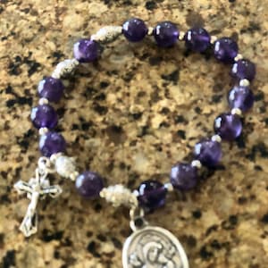 May include: A beaded bracelet featuring deep purple amethyst stones. The bracelet includes silver-toned accents, a small cross charm, and an oval pendant with a religious figure. The beads are approximately 8mm in diameter.