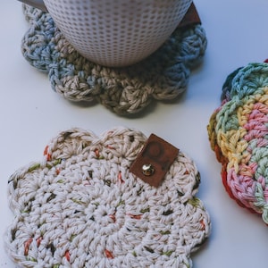 Puede incluir: Una taza de cerámica blanca llena de té descansa sobre un posavasos de ganchillo hecho a mano, blanquecino, con un borde festoneado. El posavasos tiene una etiqueta de cuero con un tachuela de metal. Otro posavasos con un patrón de arcoíris es visible.