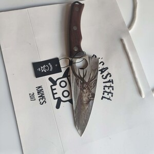 Puede incluir: Un cuchillo de cocina de acero inoxidable con mango de madera marrón. La hoja tiene un diseño de ciervo y el texto "CASTSTEEL" está impreso en la hoja. El cuchillo está empaquetado en una bolsa de papel blanca con el texto "KNIVES 2017" y un logotipo de calavera.