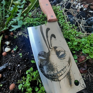 Könnte beinhalten: Ein Edelstahl-Hackmesser mit einem Holzgriff. Die Klinge zeigt eine Schwarzweiß-Illustration eines Comic-Charakters mit einem verschmitzten Grinsen.