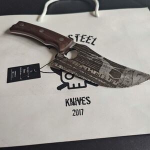 Puede incluir: Un cuchillo con mango marrón y hoja de acero inoxidable. La hoja tiene un diseño de calavera y huesos cruzados y una escena de bosque grabada. El cuchillo está sobre una bolsa de papel blanca con las palabras "STEEL KNIVES 2017" impresas en ella.