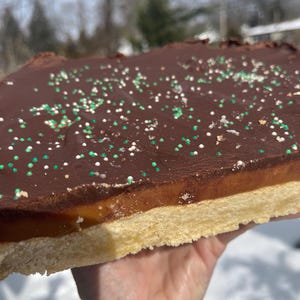May include: A hand holding a rectangular dessert bar. The bar has a shortbread base, a caramel layer, and a chocolate topping sprinkled with green and white nonpareils. The dessert is outdoors in the sunlight.