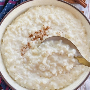 May include: A bowl of creamy rice pudding with a sprinkle of cinnamon. A gold spoon is scooping out a portion of the pudding. Cinnamon sticks and a plaid cloth are in the background. The bowl has a beige rim and a brown border.