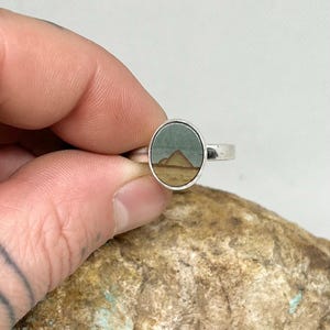May include: A silver ring featuring an oval-shaped inlay with a natural landscape pattern. The inlay displays a muted green sky above a tan desert horizon with a distinct triangular pyramid shape centered in the middle of the stone setting.