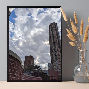 Op de afbeelding: Een ingelijste print met een stadsgezicht met hoge gebouwen onder een bewolkte lucht. De gebouwen zijn voornamelijk bruin en beige, met een bakstenen muur op de voorgrond. Een vaas met gedroogde tarwestengels staat rechts.