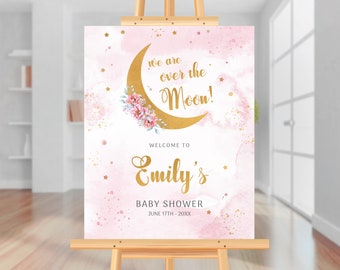 May include: Pink and gold baby shower welcome sign with a gold crescent moon and floral design. The text reads "We are over the Moon!" and "Welcome to Emily's Baby Shower June 17th - 20XX".