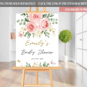 May include: A white sign with a floral design in shades of pink and green. The text "Emily's Baby Shower" is written in gold script with the date "JULY 17TH-2020" below it.