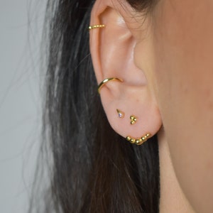 Könnte beinhalten: Nahaufnahme eines Ohrs mit mehreren goldenen Ohrringen. Die Ohrringe umfassen einen kleinen Ring, einen einzelnen Ohrstecker mit einem kleinen, klaren Stein und eine gebogene Stange mit einer Reihe kleiner goldener Kugeln.