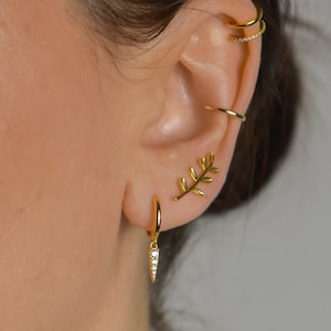 Puede incluir: Aretes de aro de oro con un pequeño colgante engastado con diamantes que cuelga de uno de los aretes. El otro arete presenta un delicado diseño de hoja de oro.