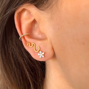 Könnte beinhalten: Nahaufnahme eines Ohrs mit drei goldenen Ohrringen. Die Ohrringe sind eine kleine Blume mit klaren Steinen, ein wellenförmiges Design und ein kleiner Ring mit klaren Steinen.