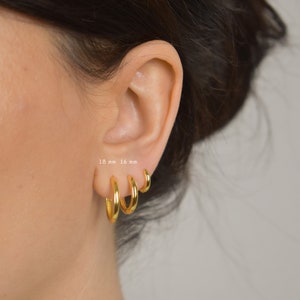 Puede incluir: Tres pendientes de aro dorados de diferentes tama&ntilde;os se muestran en una oreja. Los pendientes miden 18 mm, 16 mm y un tama&ntilde;o m&aacute;s peque&ntilde;o. El texto "18 mm 16 mm" es visible. Los aros tienen un acabado brillante y un dise&ntilde;o cl&aacute;sico.