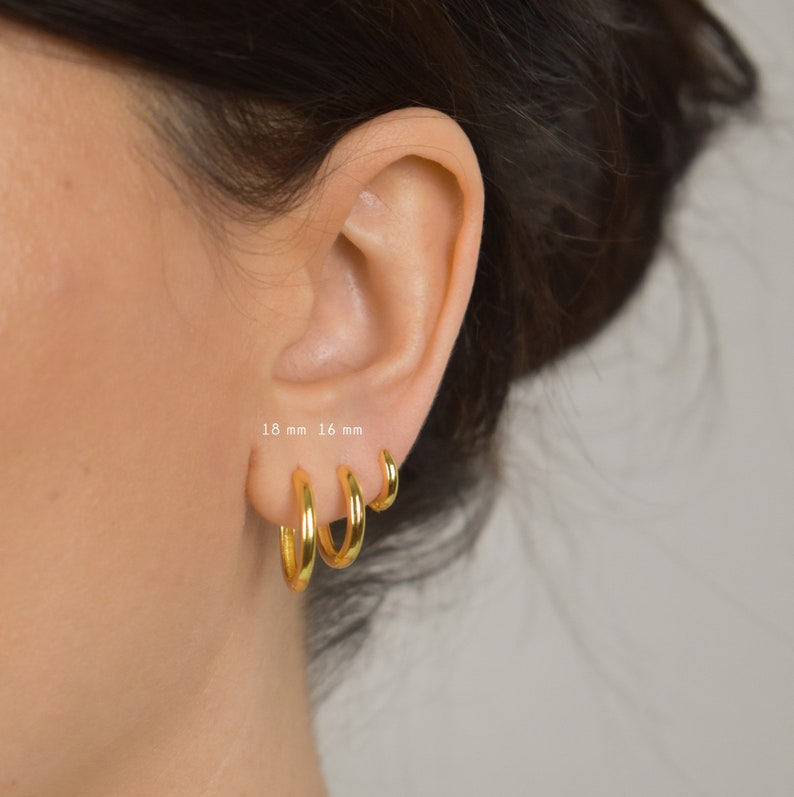 Puede incluir: Tres pendientes de aro dorados de diferentes tama&ntilde;os se muestran en una oreja. Los pendientes miden 18 mm, 16 mm y un tama&ntilde;o m&aacute;s peque&ntilde;o. El texto "18 mm 16 mm" es visible. Los aros tienen un acabado brillante y un dise&ntilde;o cl&aacute;sico.