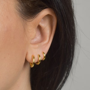 Puede incluir: Tres peque&ntilde;os pendientes de aro dorados se llevan en una oreja. Los pendientes est&aacute;n espaciados uniformemente y tienen un acabado liso y brillante. La oreja est&aacute; colocada sobre un fondo neutro, con cabello oscuro visible en el lado derecho.