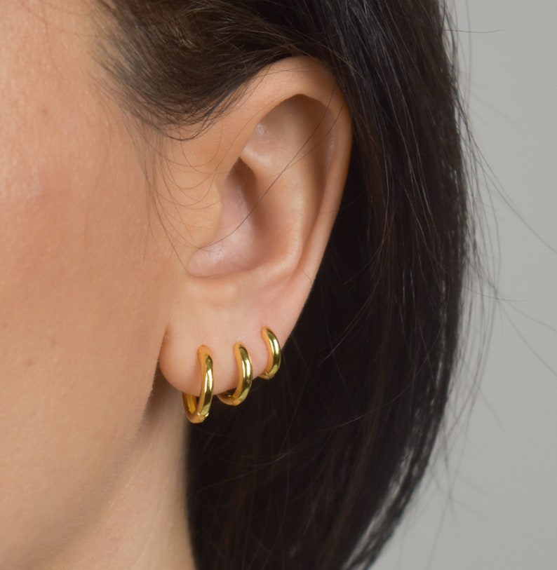 Puede incluir: Tres peque&ntilde;os pendientes de aro dorados se llevan en una oreja. Los pendientes est&aacute;n espaciados uniformemente y tienen un acabado liso y brillante. La oreja est&aacute; colocada sobre un fondo neutro, con cabello oscuro visible en el lado derecho.