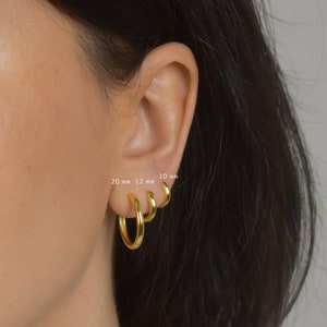 Puede incluir: Tres pendientes de aro dorados de diferentes tama&ntilde;os se muestran en una oreja. Los tama&ntilde;os est&aacute;n etiquetados como 20 mm, 12 mm y 10 mm. Los pendientes son brillantes y reflejan la luz.