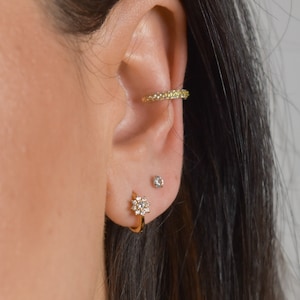 Peut inclure: Gros plan sur l'oreille d'une personne avec trois boucles d'oreilles en or. La boucle d'oreille du haut est une bande fine avec de petites pierres claires. La boucle d'oreille du milieu est un petit clou en pierre claire. La boucle d'oreille du bas est un anneau avec un motif en forme de fleur fait de pierres claires.