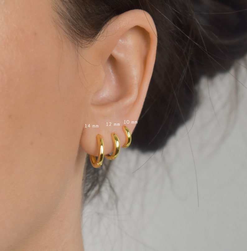 Puede incluir: Primer plano de tres pendientes de aro dorados, cada uno de un tama&ntilde;o diferente: 10 mm, 12 mm y 14 mm. Los pendientes se llevan en una oreja, mostrando su dise&ntilde;o sencillo y pulido. La imagen destaca las variaciones de tama&ntilde;o.