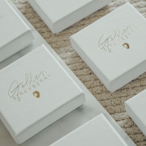 Puede incluir: Varias cajas cuadradas blancas con la inscripci&oacute;n dorada "Golden Strawberry" y un peque&ntilde;o icono de fresa. Las cajas est&aacute;n dispuestas sobre una superficie clara y texturizada, sugiriendo un contexto de venta o presentaci&oacute;n.
