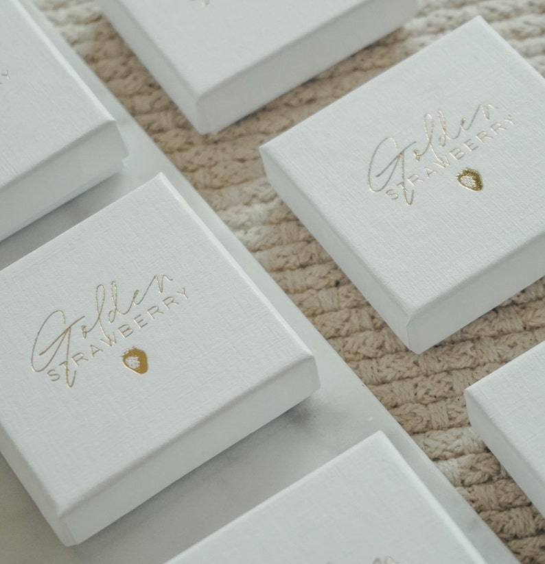 Puede incluir: Varias cajas cuadradas blancas con la inscripci&oacute;n dorada "Golden Strawberry" y un peque&ntilde;o icono de fresa. Las cajas est&aacute;n dispuestas sobre una superficie clara y texturizada, sugiriendo un contexto de venta o presentaci&oacute;n.