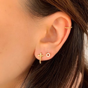 Könnte beinhalten: Nahaufnahme eines Ohres mit drei goldenen Ohrringen. Die Ohrringe sind ein kleiner Kreis, ein kleiner Kreis mit kleinen Steinen und ein Kreuz mit einer kleinen Kette.