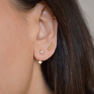 May include: Close-up of an ear adorned with three gold earrings. Two are square-shaped with clear gemstones, and the third is a linear design with small, square gemstones. The earrings are set against the backdrop of dark brown hair.