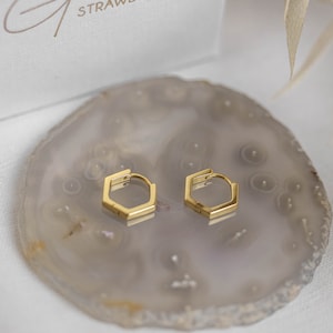 Puede incluir: Un par de pendientes de aro hexagonales dorados sobre un posavasos de ágata gris claro.