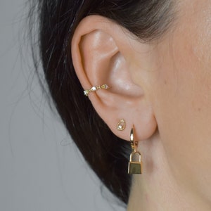 Peut inclure: Boucles d'oreilles créoles en or avec un petit charme en forme de larme et un charme en forme de cadenas suspendu au bas. Les boucles d'oreilles sont portées à l'oreille d'une personne.