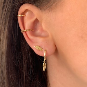 Op de afbeelding: Gouden veren oorbellen met een hoepel en een klein veren bedeltje. De oorbellen worden in de oorlel en de helix gedragen.