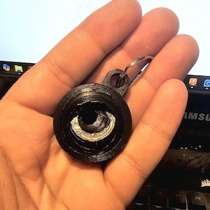 May include: A black plastic keychain with a silver metal ring. The keychain has a round, multi-layered design with a central hole.