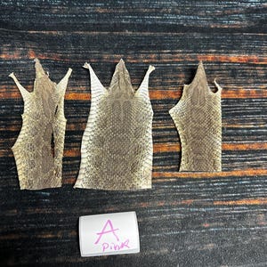 May include: Three pieces of tanned reptile skin, likely snake, with a light brown color and a scaly texture. The skin is arranged in a row on a dark wood surface. A white label with the letter "A" and the word "Pink" written in pink marker is below the skin.