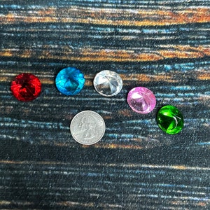 May include: Six colorful gemstones on a dark wood background. The gemstones are red, blue, clear, pink, and green. A US quarter is included for scale.