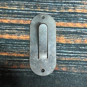 May include: A vintage, oval-shaped metal latch with a U-shaped handle. The latch has two small holes at the top and bottom. The metal has a worn, aged appearance, with signs of oxidation and wear.