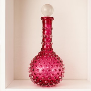 May include: A decorative, ruby-coloured glass bottle with a round, textured body and a long, beaded neck. The bottle has a clear, round stopper. The bottle is displayed on a white shelf.