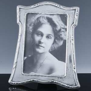 May include: A silver-toned picture frame with an ornate, curved design. The frame holds a black and white photograph of a woman. The frame has a textured surface and a decorative border. The photograph is centered within the frame.