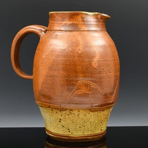 Peut inclure: Pichet en céramique fait à la main avec une grande anse et un bec verseur. Le pichet est principalement marron avec une base jaune-brun clair. La surface a un aspect texturé, suggérant un style rustique ou artisanal.