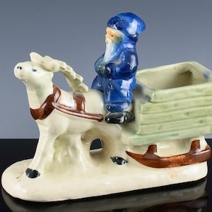May include: A ceramic figurine of a Santa Claus riding a white reindeer pulling a green wooden sled. The Santa Claus is wearing a blue hat and coat. The reindeer has brown antlers and a brown harness. The sled has a green wooden box.