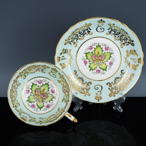 May include: A vintage teacup and saucer set. The set has a pale blue background with gold detailing and a central design of a crown, green leaf, and pink roses. The cup has a handle and the saucer is round.